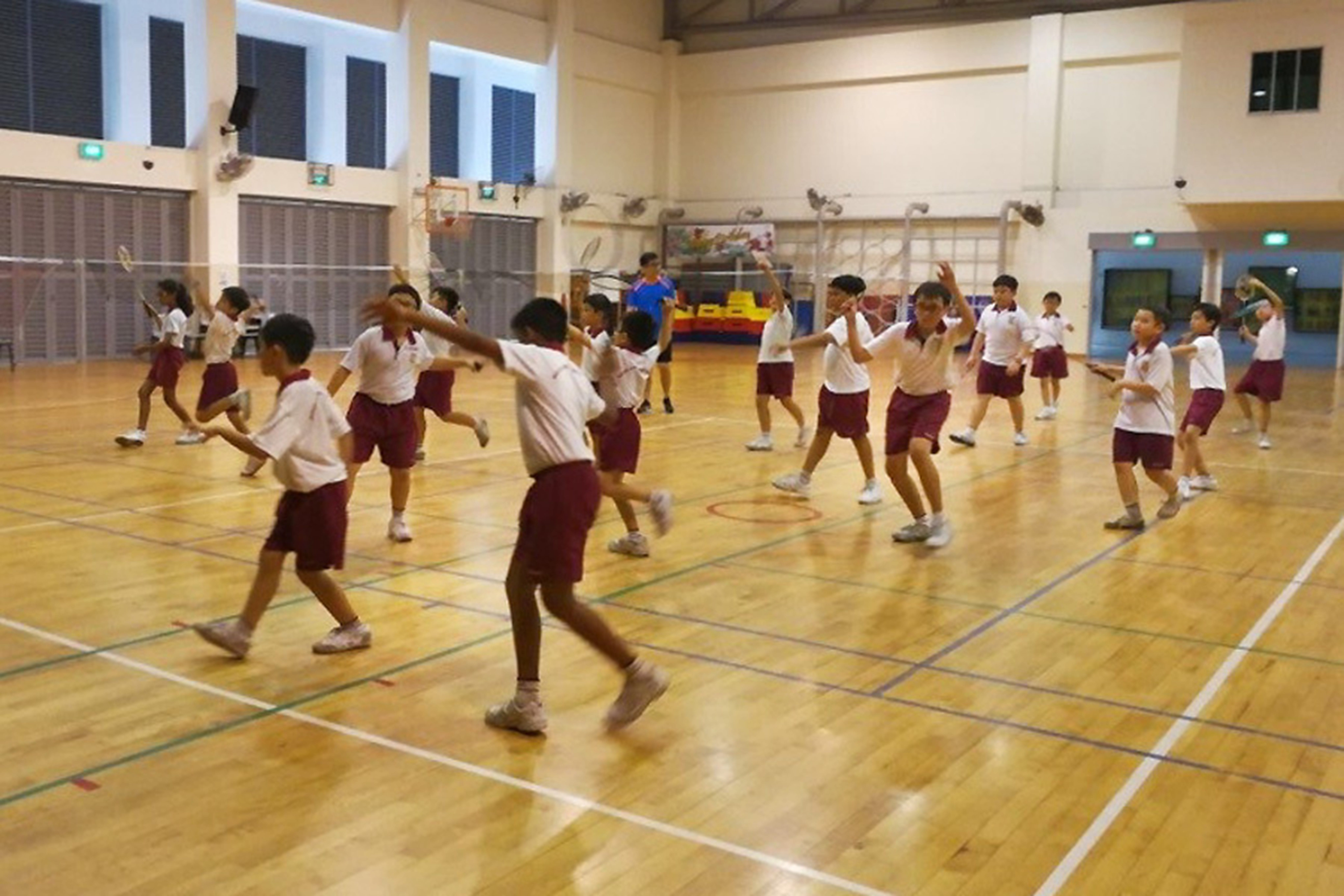 Students in white shirts and maroon shorts practice badminton in a school gym, with a coach behind them